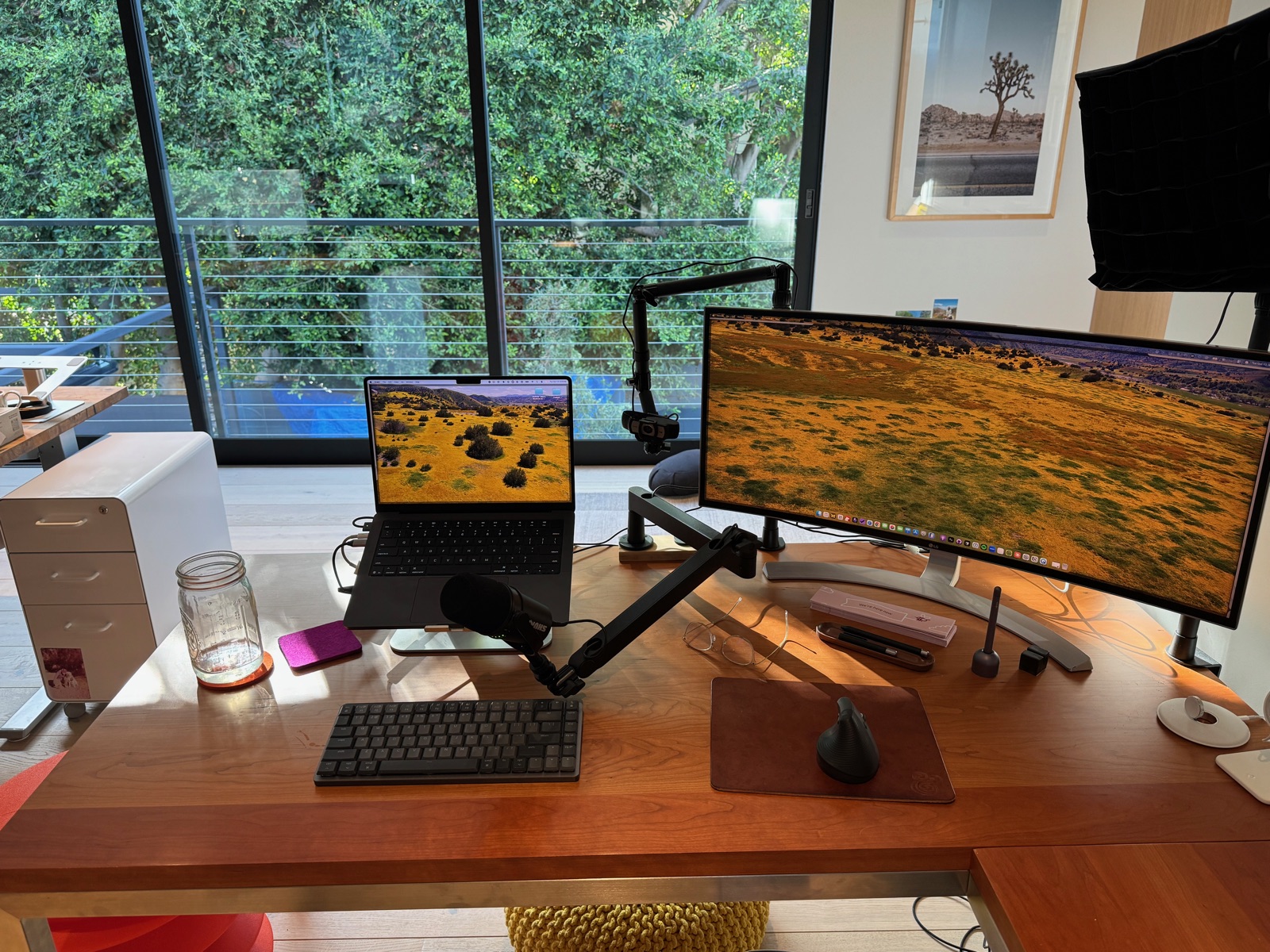 My wooden writing desk with a laptop on a stand, curved monitor, keyboard, vertical mouse, microphone, and mason jar beside large windows overlooking trees.