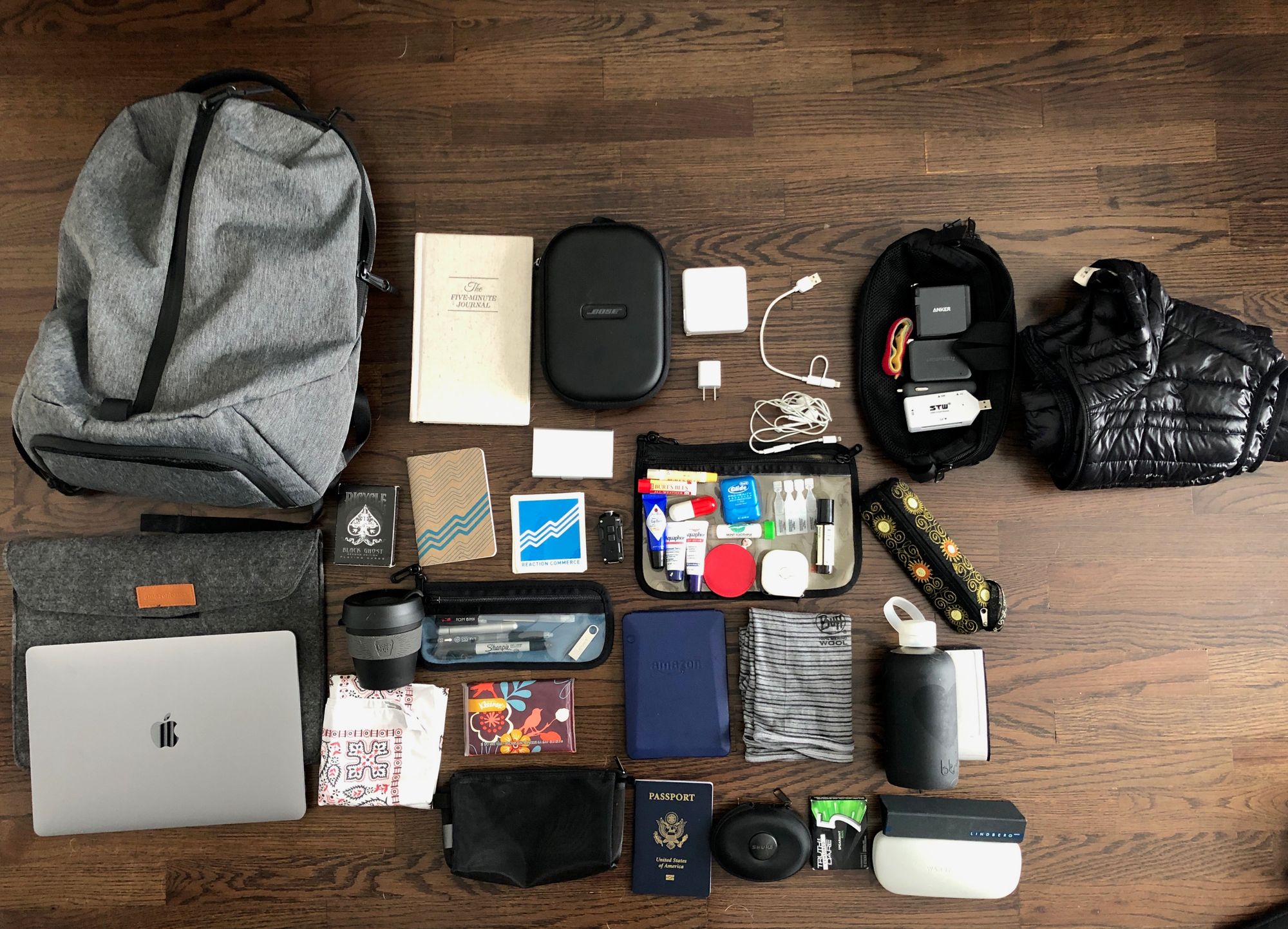 A gray backpack and its travel contents arranged on a wooden floor, including a laptop, journal, cables, toiletries, clothing, and passport